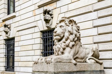 Bir binanın önündeki taş bir kaidede bir aslan heykeli duruyor. Heykel büyük ve muhteşem bir görünüme sahip. Heykelin arkasındaki bina eski ve klasik bir tasarımı var.