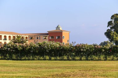 İtalya 'nın Sardunya adasında yemyeşil bir arazinin önünde yeşil çatılı büyük bir bina yer almaktadır. Bina bir çiçek çiti ve çalılarla çevrili. Gökyüzü açık ve mavi ve görünürde hiç bulut yok.