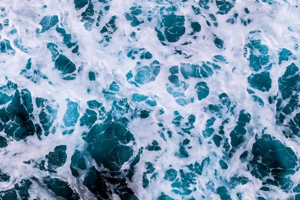 Görüntüde mavi ve beyaz desenli bir su kütlesi var. Su dalgalı ve dalgalı görünüyor, dalgalar kıyıya çarpıyor. Sahne, hareket ve enerjiden oluşur.