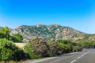 İtalya 'da Sardunya adasında, arkasında dağ olan bir yol. Yol boş ve gökyüzü temiz. Dağ ağaçlar ve çalılarla kaplı.