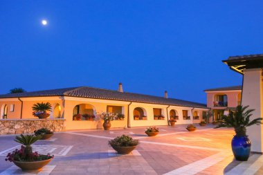 Sarı cepheli ve geniş kemerli pencereli bir bina. Binanın balkonunda beyaz bir sandalye ve beyaz bir masa var. Balkon gece aydınlanıyor.