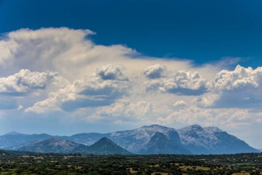 Gökyüzü mavi ve bulutlu. Bulutlar beyaz ve kabarık. Dağlar arka planda.