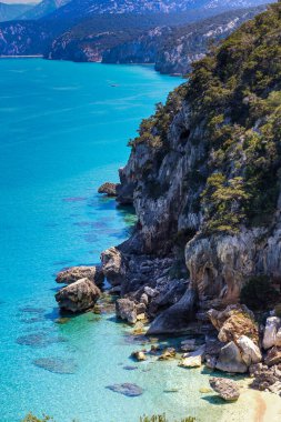 İtalya 'daki Sardunya adasında kayalık bir kıyı şeridi olan güzel mavi bir deniz. Su sakin ve berrak, ve gökyüzü parlak bir mavidir. Sahne sakin ve sakin, arkada okyanus ve dağlar var.