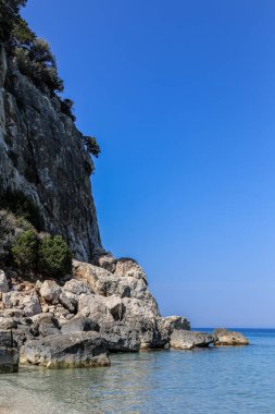 İtalya 'daki Sardunya adasının arka planında berrak mavi bir okyanusu olan kayalık bir sahil. Kumsal küçük kayalar ve deniz kabuklarıyla kaplı.