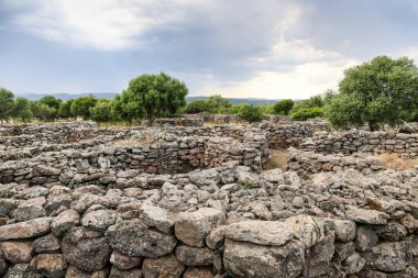 Birkaç ağacı ve bulutlu gökyüzü olan kayalık bir tarla. Sahne, İtalya 'daki Sardunya Adası' ndaki Nuraghe-Mannu Arkeolojik Bölgesi 'ndeki antik bir taş kapı binası kadar kasvetli ve ıssız.