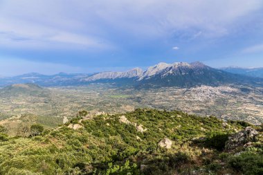 Arka planda, İtalya 'da Sardunya adasının ön planında bir kasabayla birlikte bir dağ sırası görülebilir. Kasaba yemyeşil bir manzarayla çevrili, ağaçlar ve kayalar etrafa saçılmış.