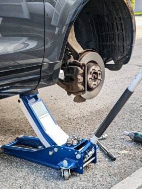 Siyah bir araba kriko tarafından kaldırılıyor. Araba bir caddede. Lastik gri ve fren disklerini görebilirsiniz..