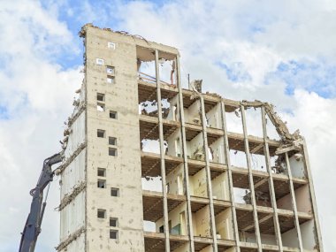 Bir vinçle bir bina yıkılıyor. Bina yüksek ve birçok penceresi var. Gökyüzü bulutlu ve güneş parlıyor
