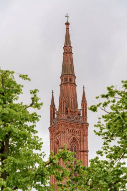 Wiesbaden Almanya 'sındaki Market Kilisesi' nin tepesinde haç olan uzun kırmızı tuğla bir kulesi. Kule ağaçlarla çevrili ve gökyüzü bulutlu.