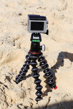 Kumlu bir sahilde, tripodun üzerinde bir kamera var. Kamera siyah ve beyaz. Kamera kullanımda değil.