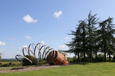 Büyük bir turuncu fıçı çimenli bir alanda yerde oturuyor. Namlu metal bir spiral ile çevrili ve arka planda ağaçlar var. Sahnenin kırsal ve doğal bir havası var.