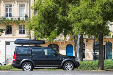 Çatısı olan siyah bir araba bir binanın önünde park edilmiş. Araba arka planda ağaç olan bir caddede park halinde.