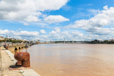 Jacques Chaban Delmas Köprüsü, arka planda Fransa 'da bir Bordeaux şehri bulunan Garonne Nehri' ni kapsar. Gökyüzü çoğunlukla bulutlu, ama güneş bulutların arasından parlıyor.