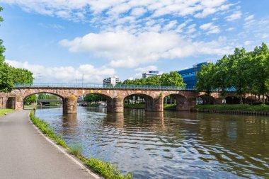Eski bir köprü, Saarbrucken Almanya 'da arka planda bir şehir manzaralı bir Saar nehri boyunca uzanır. Köprü taştan yapılmıştır ve mavi ve beyaz bir renk düzenine sahiptir. Su sakin ve gökyüzü berrak.