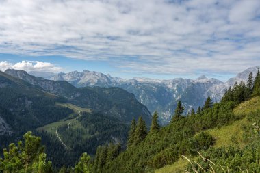 Almanya 'da, kısmen ormanla kaplı gri dağları olan güzel bir dağ manzarası. Gökyüzü mavi ve bulutlu. Dağların arasından geçen ince, dolambaçlı bir yol..