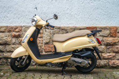 Scooter evin taş duvarının yanına park edilmiş. Mobilet bej renkli ve bej koltuklu. Direksiyon sola döndü..