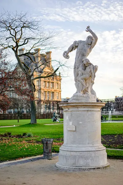 Bir demet üzüm tutan bir adam heykeli Paris 'te bir binanın önünde duruyor. Heykel beyaz ve bina kahverengidir. Heykelle manzara huzurlu ve huzurlu.