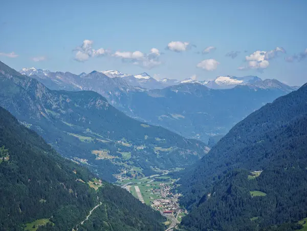 Aşağıdaki vadide bir kasabası olan bir Alpler dağ sırası. Kasaba yeşil tepeler ve ağaçlarla çevrilidir. Gökyüzü açık ve mavidir, ve güneş parlıyor. Huzurlu ve huzurlu bir sahne.