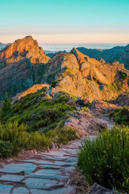 A scenic view of a mountain trail with hikers, surrounded by lush greenery, illuminated by golden sunlight, Pico do Arieiro, Madeira. clipart