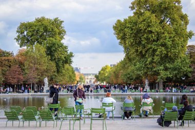 People relax and enjoy the peaceful atmosphere by the pond in a park in Paris, with green chairs and trees lining the path. clipart