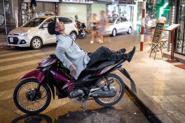 A man relaxes on his motorcycle on a bustling street at night in Vietnam, capturing the essence of urban life with vibrant city lights.