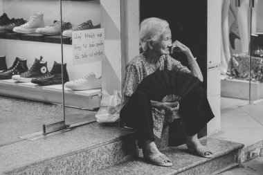 An elderly woman seated on the steps of a store with a fan in hand, takes a moment of rest while the hustle of the street continues around her.