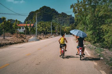 Two boys ride bicycles on a rural road in Vietnam, one holding a large umbrella for shade. The scene captures a sunny day amidst scenic mountain view. clipart