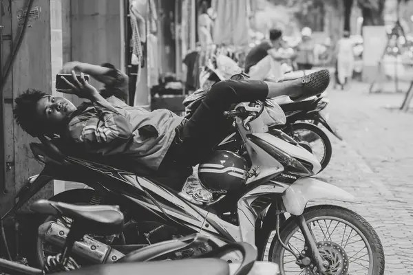 Genç bir adam motosikletinin üstünde rahatlıyor, telefonuyla kayıyor, Hanoi 'nin işlek caddesinde, siyah beyaz bir şekilde huzurlu bir anı yakalıyor..