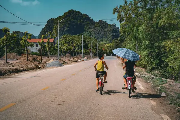 Vietnam 'da kırsal bir yolda bisiklete binen iki çocuk, biri gölge için büyük bir şemsiye tutuyor. Manzaralı dağ manzarası arasında güneşli bir günü yakalar.