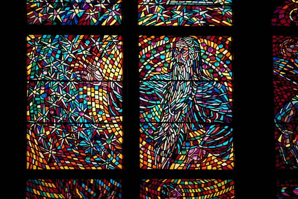 A close-up of a colorful stained glass window, depicting abstract religious imagery with vibrant hues of red, blue, yellow, and purple, in St Vitus Church in Prague, Czechia.