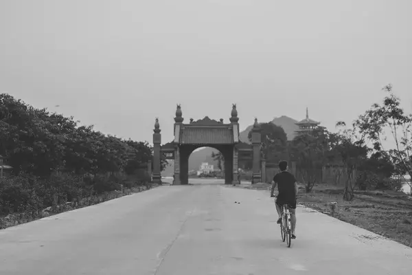Yalnız bir bisikletçi, sakin ve boş bir yol sahnesinde geleneksel bir Vietnam kapısına doğru gidiyor. Siyah-beyaz tonlar huzurlu ortamı vurguluyor..