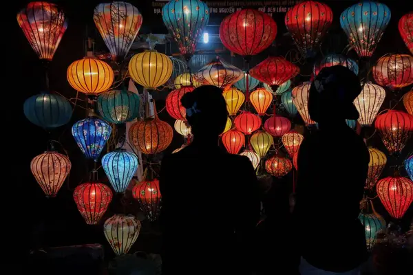 İki kişinin siluetleri Hoi An, Vietnam 'da geceleri göz kamaştırıcı bir görsel karşıtlık yaratan renkli fenerlere hayranlık duyuyor..