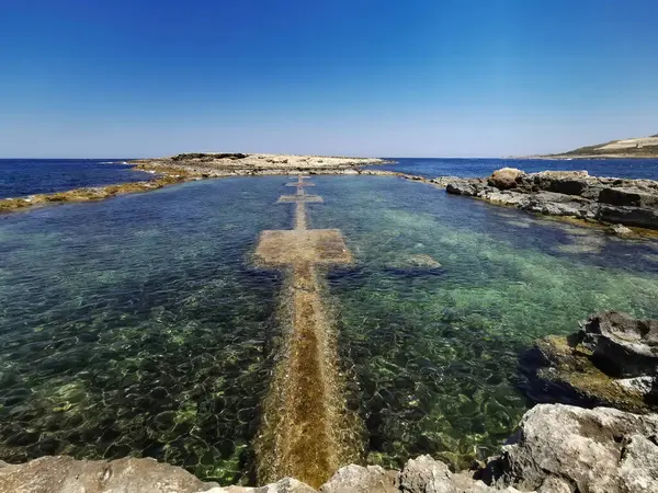 Parlak bir gökyüzü altında kayalık bir sahil boyunca mavi-yeşil suyu olan temiz doğal kaya havuzu, Malta.