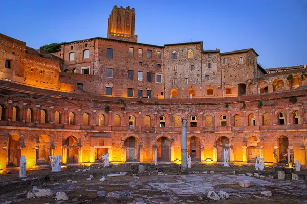 The ancient Roman ruins of Trajan's Markets illuminated at dusk, featuring sculptures and a column, with the surrounding buildings in the background.