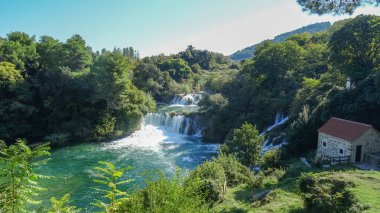 Hırvatistan 'ın Krka Ulusal Parkı' ndaki Skradinski Buk şelalesinin panoramik manzarası, geleneksel taş ev, yeşil orman ve parlak gökyüzünün altındaki temiz zümrüt nehri.
