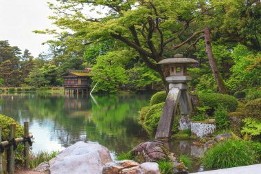 Kenrokuen Garden, Kanazawa 'da yemyeşil bir gölet ve arka planda ahşap bir çay evi olan geleneksel Japon taş feneri..