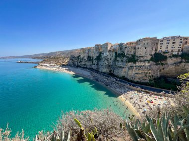 İtalya 'daki Tropea plajının panoramik manzarası, turkuaz deniz, sahile gidenler ve kıyı kayalıklarının tepesine tünemiş tarihi binalar..
