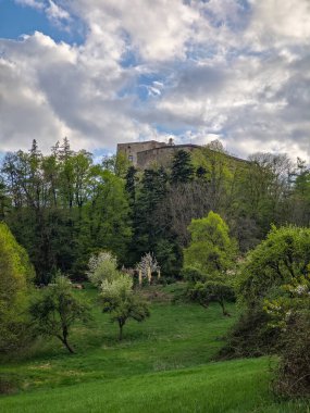 Buchlov Şatosu yemyeşil bahar ağaçları ve çiçek açan meyve bahçesinin üzerinde yükselir, parçalı bulutlu bir gökyüzünün altındaki çayırdan fotoğraflanır..