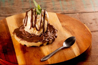 Çikolatalı waffle, fındıklı krema ve önünde tatlı kaşığı olan yuvarlak tahta bir tahtada bir kepçe vanilyalı dondurma.