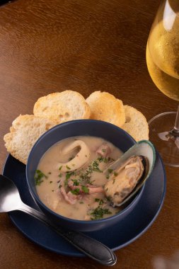 Bir kase deniz ürünleri çorbası, kızarmış Yeni Zelanda midyesi ve ahşap masada yeşil soğan. En üstteki portrede beyaz şarap kadehiyle üst manzara eşleşiyor.