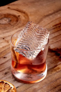 Profesyonel dekore edilmiş, şeffaf dalgalı buz küpünü, portakal renkli negroni kokteyline yakın planda ahşap masaya koymak.
