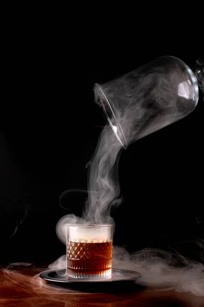 removing the drink smoker from a glass of smoked whiskey with smoke on a wooden table, black background