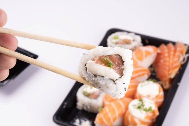 Elinde yemek çubuklarıyla bir parça suşi ve arka planda bir tepsi oryantal yiyecek vardı.