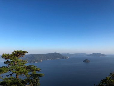 This photo is from the top of Mount Misen in Hiroshima, Japan. After hiking through the forest you reach an overlook of the Seto Inland Sea. The moon is visible in the sky. clipart