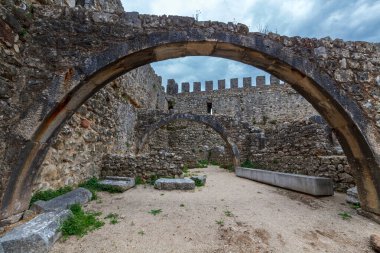 Yıpranmış duvarlar ve siperlerle çevrili kale avlusundaki antik taş kemerler, bulutlu bir gökyüzü altında ortaçağ mimarisinin dayanıklılığını ve zanaatkarlığını gözler önüne seriyor.