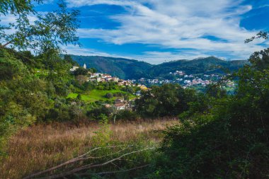 A scenic view of a small town nestled in a valley among rolling hills and lush vegetation under a blue sky