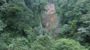Dağlarda ağır ve ağaçlarla kaplı bir şelalenin insansız hava aracı videosu, insansız hava aracı ilerliyor ve kamera şelaleye bakıyor.