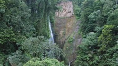 Şelalenin uzaktan çekilmiş videosu ağır ve ağaçlarla kaplanmış gibi görünüyor. Drone sağa doğru hareket ediyor ve kamera şelaleye bakıyor.