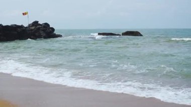 Okyanus sahilinde güneşli güzel bir gün. Arka planda kaya ve Sri Lanka bayrağı var.