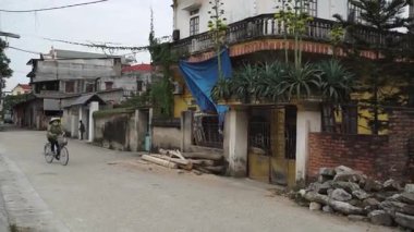 Konik şapkalı bir kadın sessiz bir Vietnam köyü caddesinde bisiklet sürüyor.